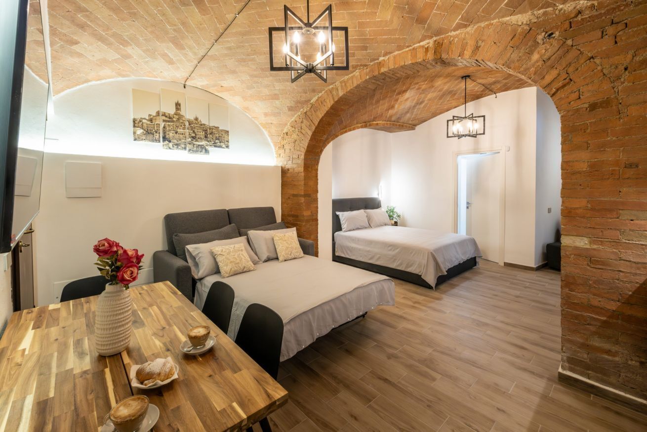 Ein Zimmer mit Holztisch, einem Bett und einem weiteren Bett im Schlafbereich, gestaltet im Loft-Stil mit freiliegenden Ziegelwänden und Blick auf die Stadt.