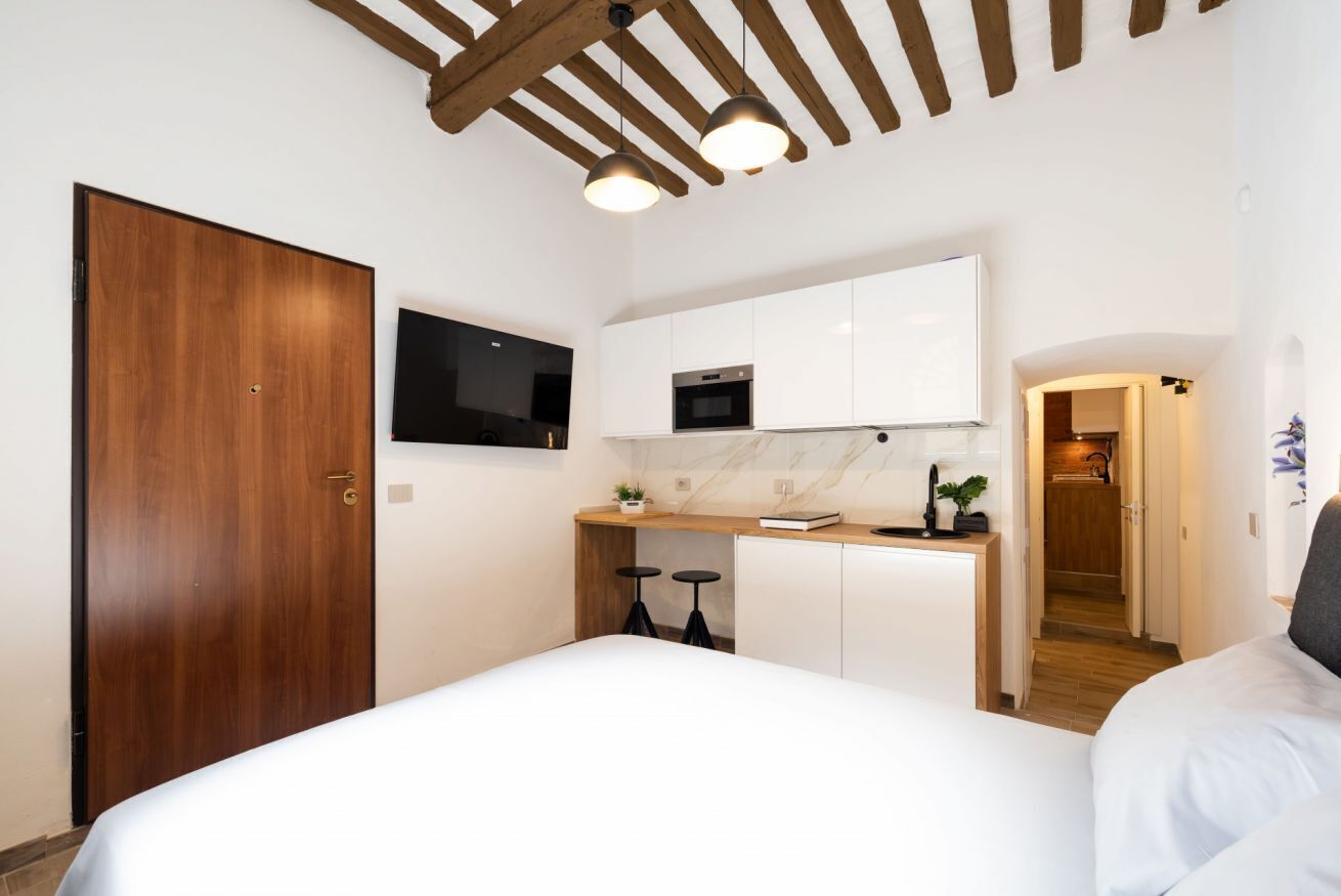 Una habitación compacta con puerta de madera, cama, kitchenette pequeño con microondas y televisión montada en la pared.