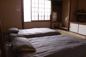 Ein Zimmer mit zwei Betten, einem Fenster, einem Ventilator und einem Regal mit einer kleinen Tierfigur.