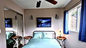 Un dormitorio con cama, dos mesitas de noche, ventilador de techo, ventana con cortinas y dos luces fijadas a la pared.