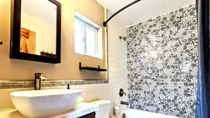 Un baño con lavamanos sobre encimera, inodoro, ducha con azulejos decorativos, espejo y ventana.