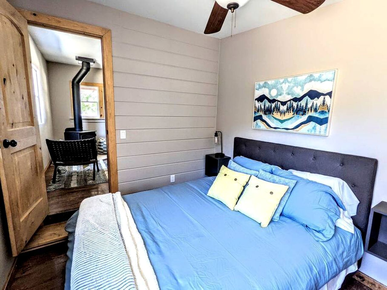 Un dormitorio con cama, mesita de noche, un cuadro y una puerta que da a otra habitación.