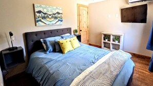 Quarto com cama, ventilador de teto, TV fixada na parede e uma pequena estante com plantas.