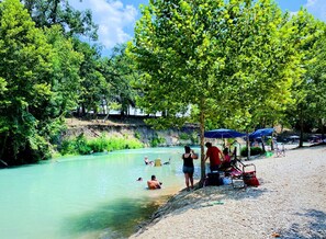 Margem de rio com pessoas nadando e relaxando, cercada por árvores e guarda-sóis.