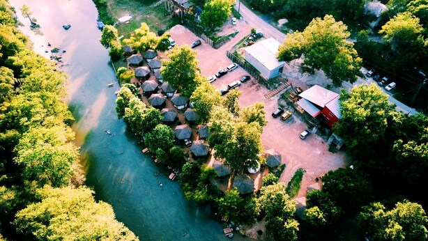 An aerial view of a riverside property with a cluster of thatched-roof huts, a main building, and a parking area.