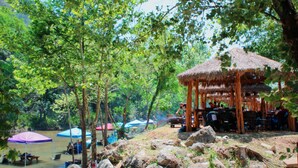 A riverside setting with a thatched-roof pavilion, picnic tables, and umbrellas.