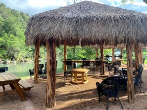 Un pavillon à toit de chaume doté d’un espace extérieur avec des sièges, une table de pique-nique et une vue sur un lac.
