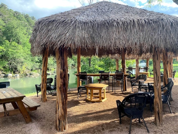 A thatched-roof pavilion with outdoor seating, a picnic table, and a view of a lake.