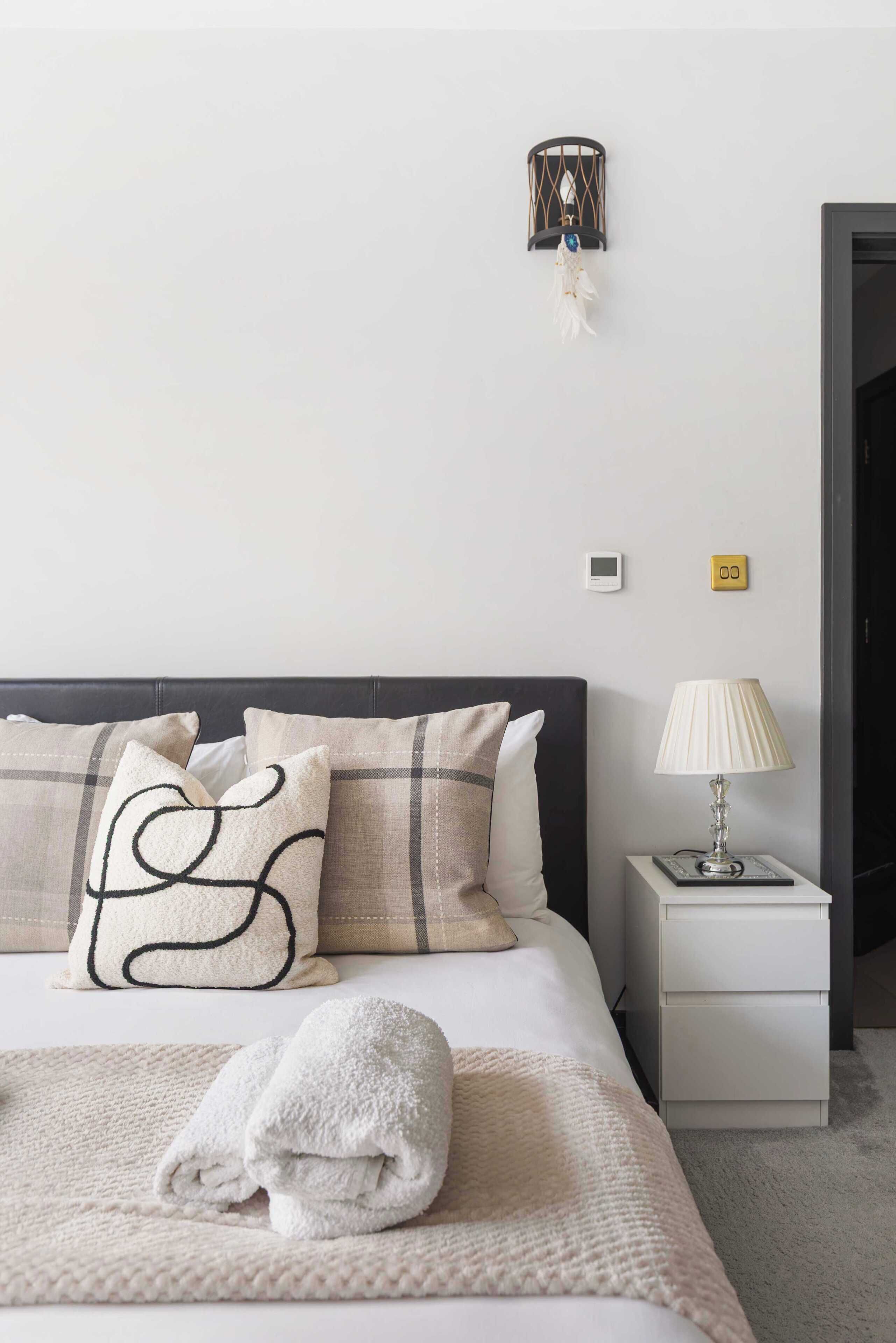 Een netjes opgemaakt bed met een witte deken en een opgerolde handdoek, een bedlamp, een klein wandlampje en een digitale thermostaat aan de muur.