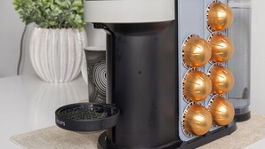 Een koffieapparaat met een waterreservoir, een houder voor koffiecups en een plantje op de achtergrond.