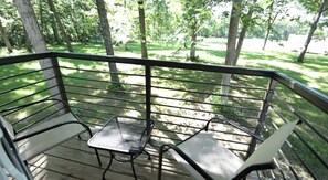 Terraza con dos sillas de metal y una mesita pequeña, rodeada por una barandilla metálica, con vistas a un área verde con árboles.