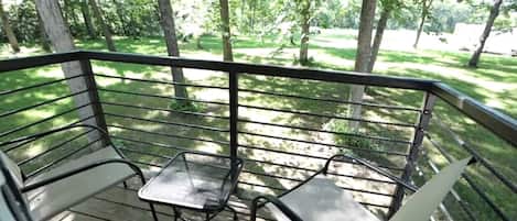 Terraza con dos sillas de metal y una mesita pequeña, rodeada por una barandilla metálica, con vistas a un área verde con árboles.