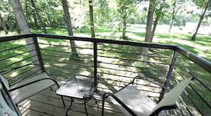 A deck with two metal chairs and a small table, surrounded by a metal railing, overlooking a grassy area with trees.