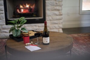 A round wooden table with a bottle of Meomi Pinot Noir, two glasses of wine, a red mug, dice, and a paper on a coaster. - Twisted Leaf Luxury Cabin | Hot Tub | Wooded Walking Trail (Broken Bow)