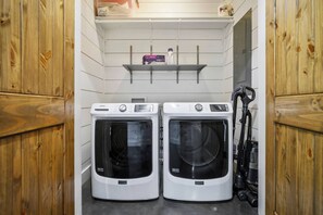 Un cuarto de lavandería con dos lavadoras de carga frontal, un estante arriba y una aspiradora a la derecha.