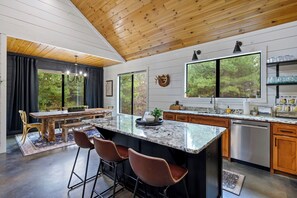 A modern kitchen with a large island, bar stools, and a dining area with a table and chairs. - Twisted Leaf Luxury Cabin | Hot Tub | Wooded Walking Trail (Broken Bow)