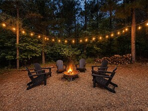 Un área con fogata, sillas Adirondack e iluminacion de luces colgantes en un entorno boscoso.