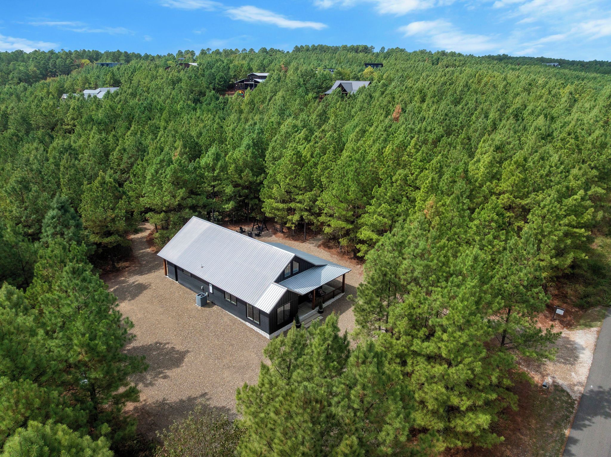 A modern cabin surrounded by dense forest, with a gravel driveway and a covered porch.