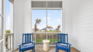 Un balcón con dos sillas azules, una mesa blanca pequeña y un ventanal con vistas a palmeras y un edificio.