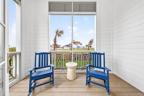 Ein Balkon mit zwei blauen Stühlen, einem kleinen weißen Tisch und einem großen Fenster mit Blick auf Palmen und ein Gebäude.