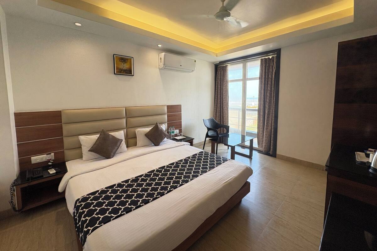 A hotel room with a large bed, a ceiling fan, an air conditioning unit, a small table with a chair, and a window with curtains.