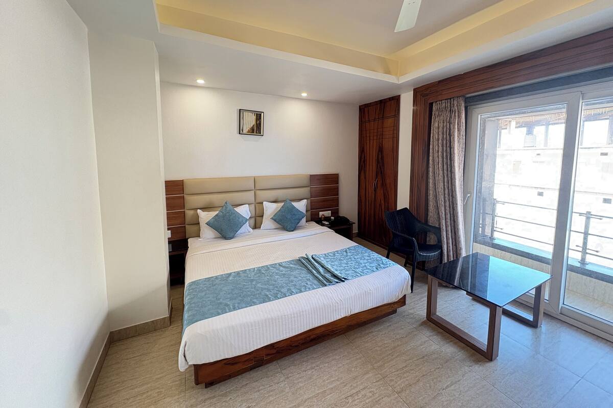 A hotel room with a bed, a desk, a chair, a ceiling fan, a window with curtains, and a door.