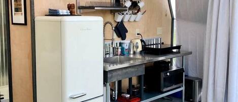 Una cocina compacta con un refrigerador blanco, un lavamanos y una estufa. Hay repisas sobre la encimera con platos y vasos. Se ve una silla negra al frente.