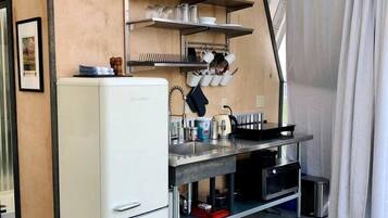 Une cuisine de taille réduite avec un réfrigérateur blanc, un évier et une cuisinière. Des étagères au-dessus du plan de travail servent à ranger la vaisselle et les verres. On aperçoit une chaise noire au premier plan.
