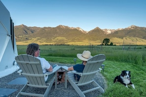 Dos personas sentadas a una mesa con un perro, con vista a un paisaje montañoso.