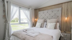 A bedroom with a large bed, a wooden headboard, a bedside table, and a window with curtains. - Swan Haven (Grange-over-Sands)