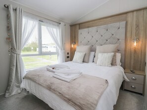 A bedroom with a large bed, a wooden headboard, a bedside table, and a window with curtains. - Swan Haven (Grange-over-Sands)