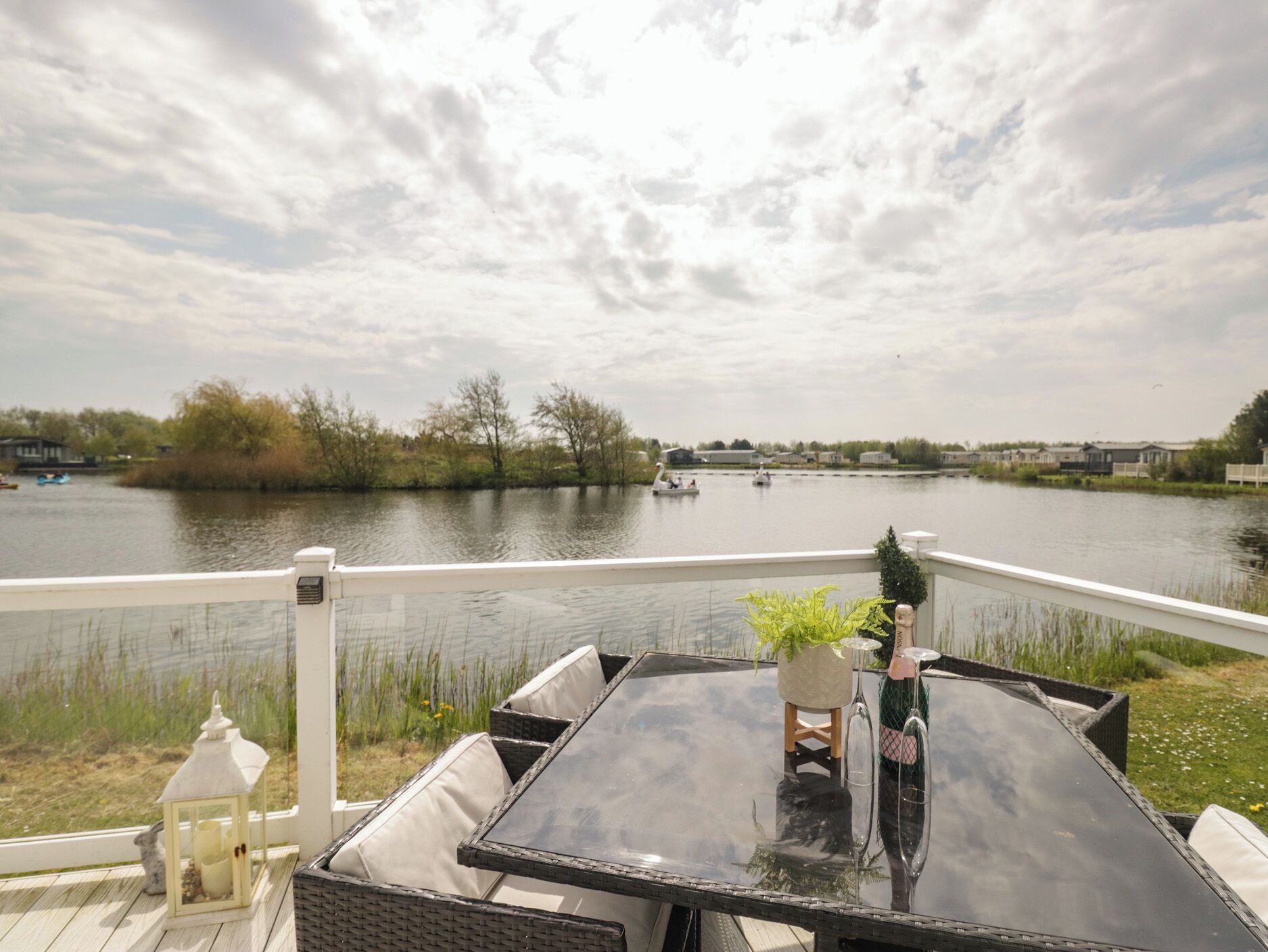 A deck with a glass table and wicker chairs, overlooking a calm lake with boats and houses in the background.