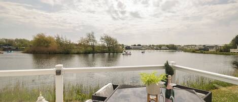 A deck with a glass table and wicker chairs, overlooking a calm lake with boats and houses in the background.