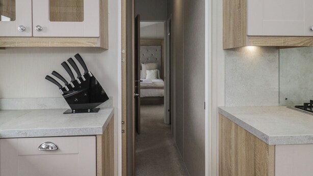 A narrow hallway with a kitchen on one side and a bedroom on the other, featuring a bed with a tufted headboard and a bedside table. - Swan Haven (Grange-over-Sands)