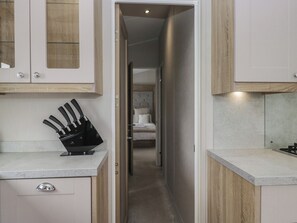 A narrow hallway with a kitchen on one side and a bedroom on the other, featuring a bed with a tufted headboard and a bedside table. - Swan Haven (Grange-over-Sands)