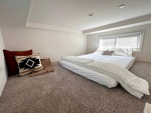 Une chambre à coucher avec un lit, une fenêtre avec des stores, un sol recouvert d’une moquette et un oreiller rouge.