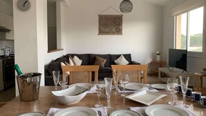 Un comedor con una mesa dispuesta para cuatro personas, un sofá, un televisor y una cocina al fondo.