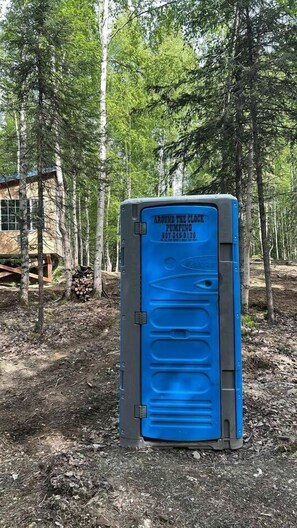 Un baño portátil con puerta azul, identificado con el rótulo 'Around the Clock Pumping', en una zona boscosa.