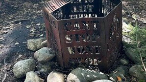 Un foyer métallique, sur lequel sont gravés les mots « LE CAMPING, ÇA NE TARIT PAS L’ENVIE », est entouré de rochers et se trouve près d’une table de pique-nique, dans un espace boisé.