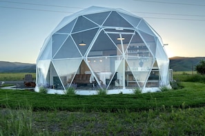 Eine geodätische Kuppelstruktur mit sichtbarem Innenraum, einschließlich Tisch und Stühlen, umgeben von einem grasbewachsenen Feld und Hügeln im Hintergrund.