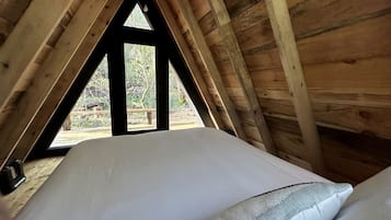 A bedroom with a sloped ceiling, a bed with white bedding, and a view through a triangular window.