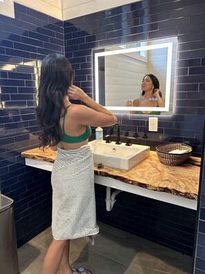 A person standing in a bathroom with a mirror, sink, and dark blue tiled walls. - Secluded Off-Grid Cabin Retreat with Fire Pit & Sauna in Zirconia, North Carolina (Zirconia)