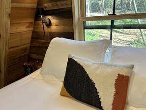A bed with white linens and a single decorative pillow in a cabin-style room with wooden walls and a window. - Secluded Off-Grid Cabin Retreat with Fire Pit & Sauna in Zirconia, North Carolina (Zirconia)