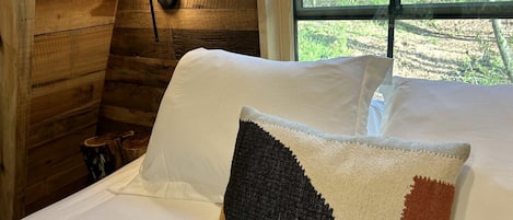 Una cama con sábanas blancas y una almohada decorativa en una habitación estilo cabaña con paredes de madera y una ventana.