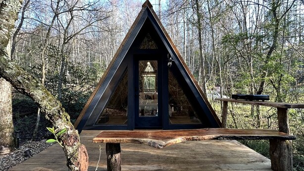 Eine Holzterrasse mit einer dreieckigen Holzkonstruktion, möglicherweise ein Pavillon oder eine Schutzhütte, umgeben von Bäumen.