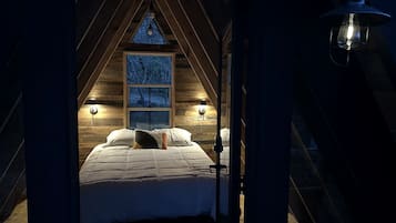 A cozy attic bedroom with a sloped ceiling, a large window with a view, and two wall-mounted lamps.