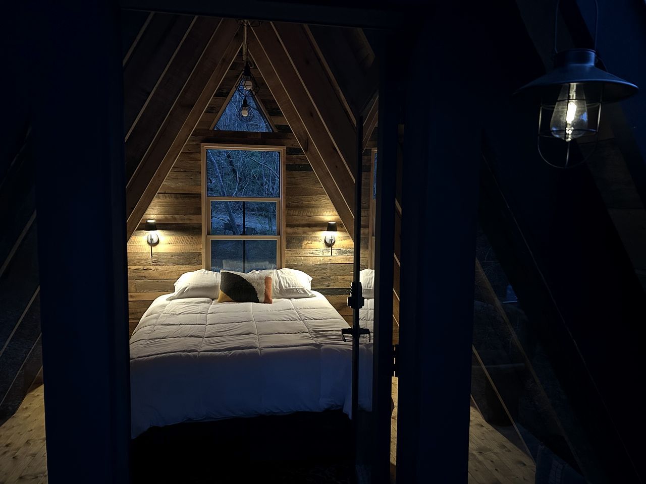 A cozy attic bedroom with a sloped ceiling, a large window with a view, and two wall-mounted lamps.
