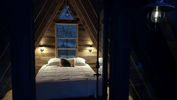 A cozy attic bedroom with a sloped ceiling, a large window with a view, and two wall-mounted lamps.