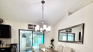 A dining area with a long table, chairs, a chandelier, a refrigerator, and a mirror on the wall.