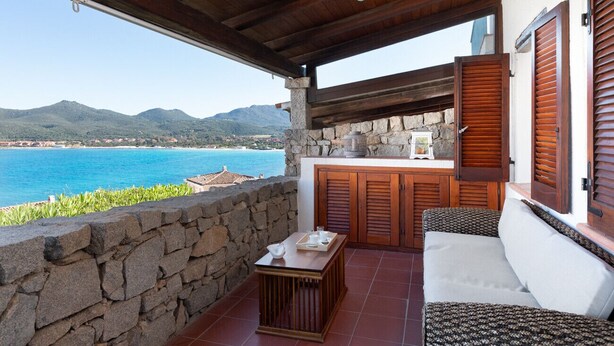 Ein Balkon mit Steinmauer, Holzmöbeln und Blick auf Meer und Berge.